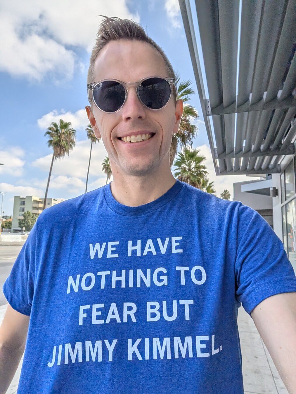 Patrick is smiling, taking a selfie outside on a sunny day. He's wearing a blue t-shirt with the words "WE HAVE NOTHING TO FEAR BUT JIMMY KIMMEL" printed on it in white letters. He's wearing sunglasses and standing in front of palm trees and a street.