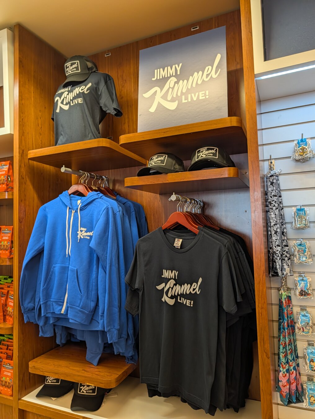 A store display featuring official merchandise for the television show Jimmy Kimmel Live! The display is made of wood and has several shelves. On the top shelf, a dark gray T-shirt with the show's logo is on a mannequin head, and a matching baseball cap is on a shelf next to it. On the middle shelf, several blue zip-up hoodies with the show's logo are hanging on a rack. Below them, on the same shelf, are two black baseball caps with the logo. On the bottom shelf, a stack of dark gray T-shirts with the show's logo is hanging on a rack.