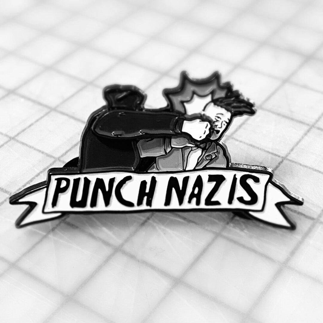 Anamel pin, were a human figure all dress in black with balaclava, is punching in the face a neonazi. Underneat says: “Punch Nazis”.