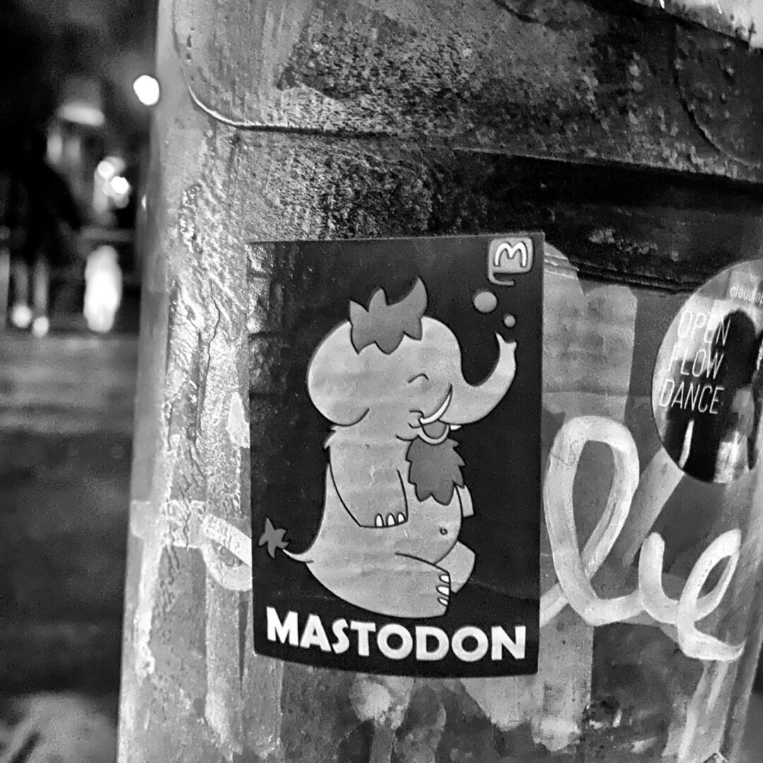 A Mastodon sticker on a public post; it contains an illustration of a Mastodon smiling while exaling an M letter.