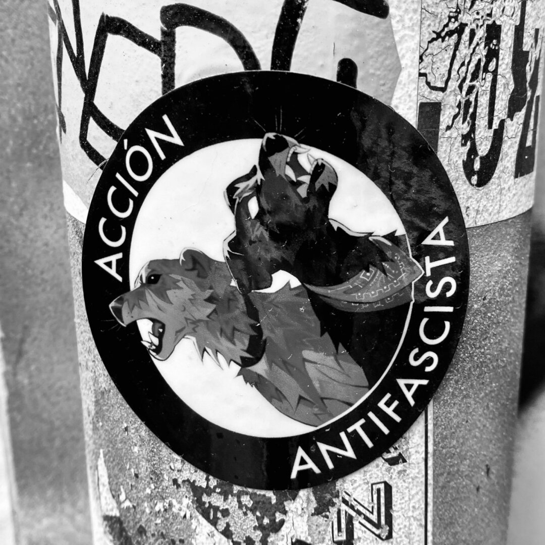 Sticker places over a post that reads: “Antifascist Action” in Spanish. The sticker contains two illustrated dogs barking at fascists.