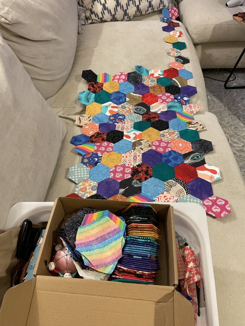 A smallish rectangle made of lots of fabric hexagons sewn together, with one edge of the rectangle extended out about twice as far as the rest of it. That edge is the full target length of the blanket. In the foreground is a cardboard box full of fabric pieces and hexagons ready to attach to the blanket.