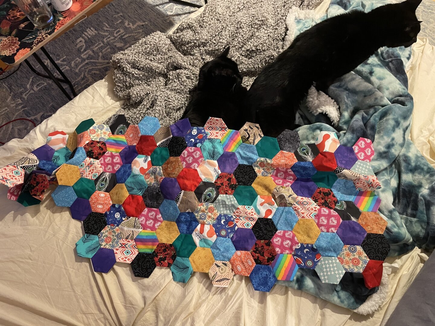 Two black cats on a pile of fuzzy blankets, underneath a sizeable quilt top made out of colorful fabric in the shape of hexagons.