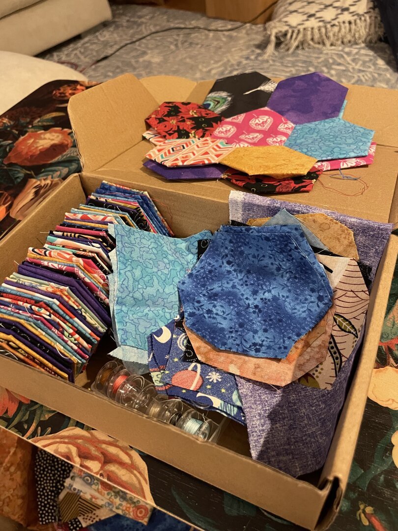 A cardboard box large enough for three rows of hexagons, currently holding one row of roughly 60 hexagons, some scrap fabric, and two pieces where I've already taken one hexagon and sewed other hexagons on each side.