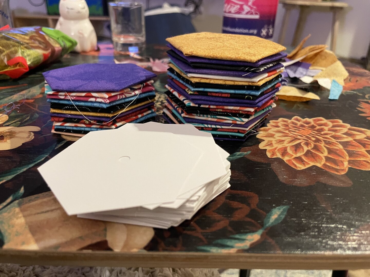 Two stacks of fabric in many colors, shaped like hexagons, with a stack of hexagon templates waiting to have fabric wrapped around them.