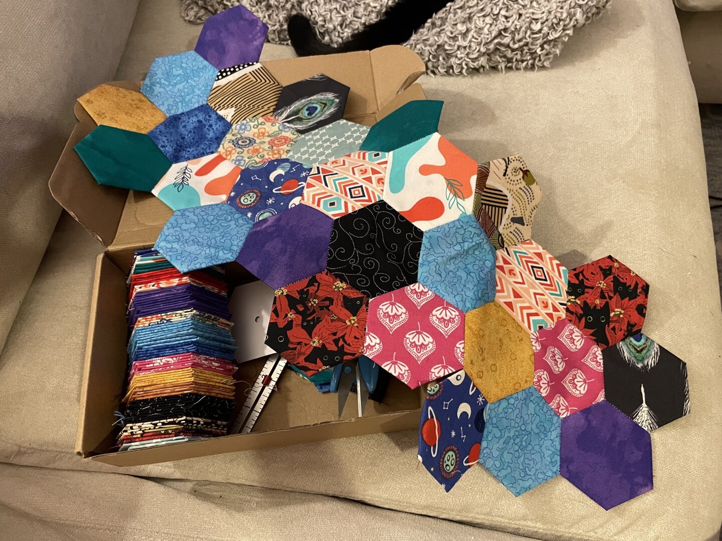 Colorful fabric hexagons arranged by color in a cardboard box, with three pieced rows of hexagons draped over the box.