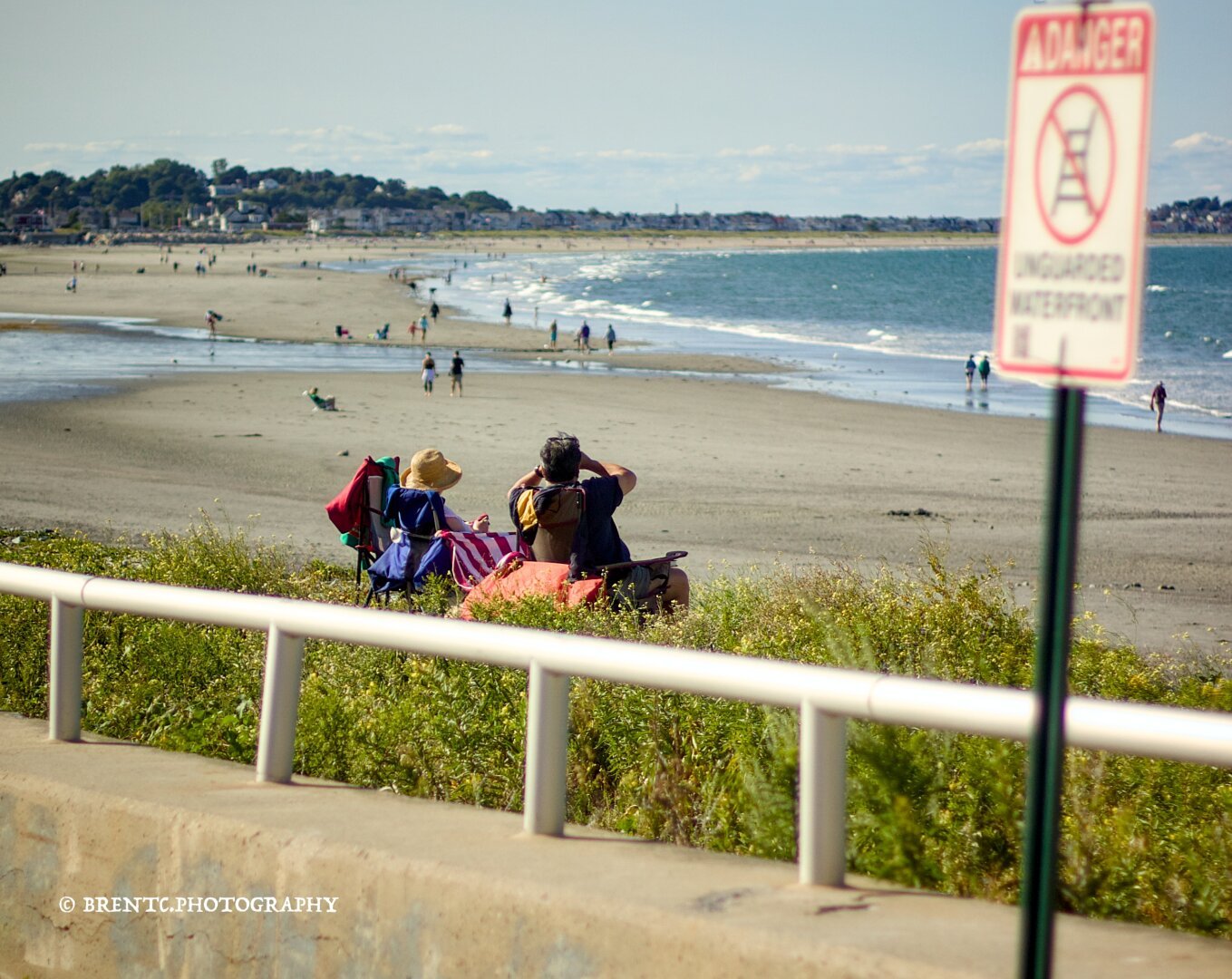 Two people on a beach in chairs watching other beachgoers through binoculars with their backs to the camera.