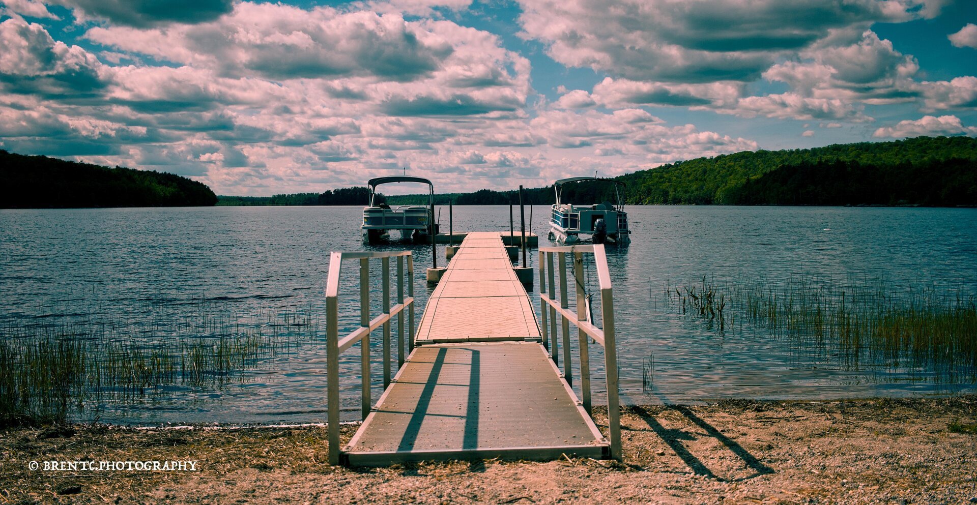 Looking down the boat dock with a boat on each side at the end. Fluffy clouds in the sky over tree-covered hills in Maine.