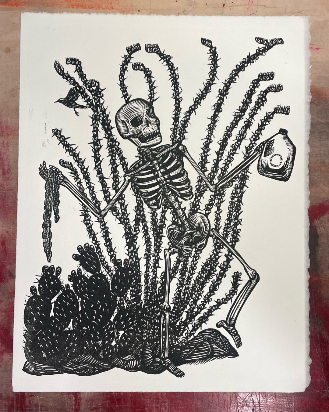 A skeleton stands infront of an ocotillo and prickly pear holding chains and jug of water.