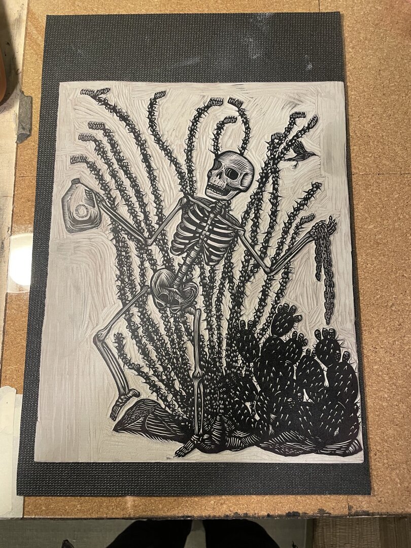 An inked linocut carving of a skeleton in front of an ocotillo holding a water jug and a chain.