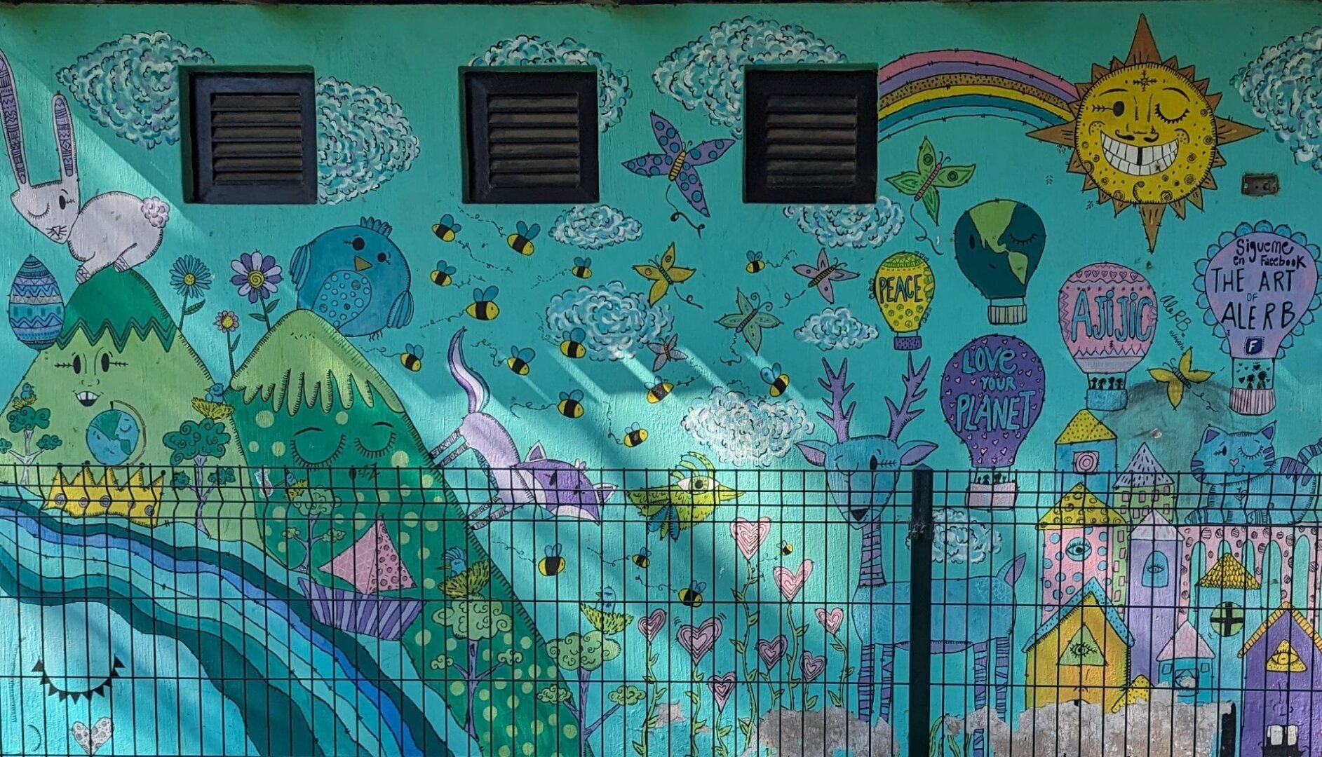 A mural of mainly blues and greens with other colours depicting mountains, a river, wildlife, and messages of love and peace on the outer wall of a small public restroom building.