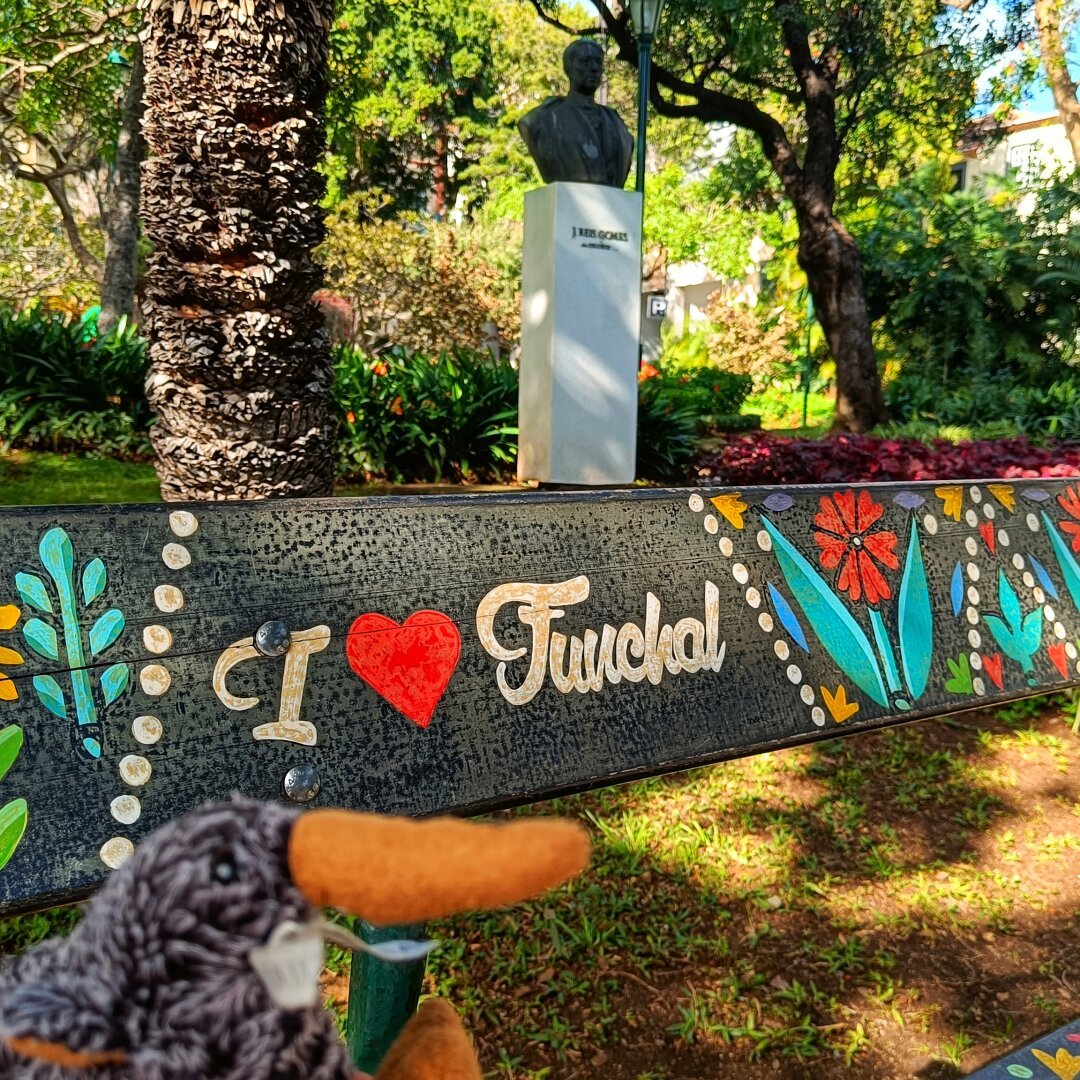 a picture of a plush kiwi in the bottom left corner. the main subject is a painted bench with flowers and "I  ❤️ Funchal" with a bust on a pedestal and green trees and plants in the background