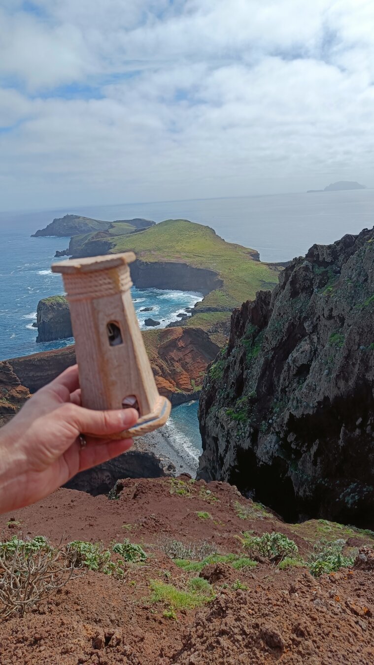 a hand holding a small hollow wooden lighthouse in front of a great view of a rocky and steep peninsula
