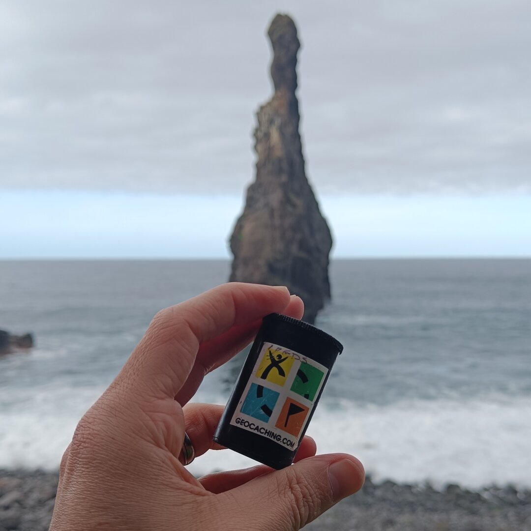 A hand holding a film canister with the geocaching logo. in the background a volcanic rock in the ocean