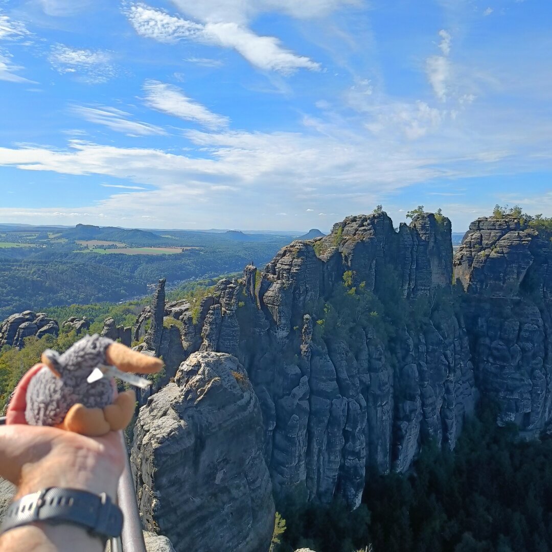 A view of the Schrammsteinfelsen, famous sandstone boulders in Saxony, Germany. In the lower left, there is a plushy kiwi, the travel mascot.
