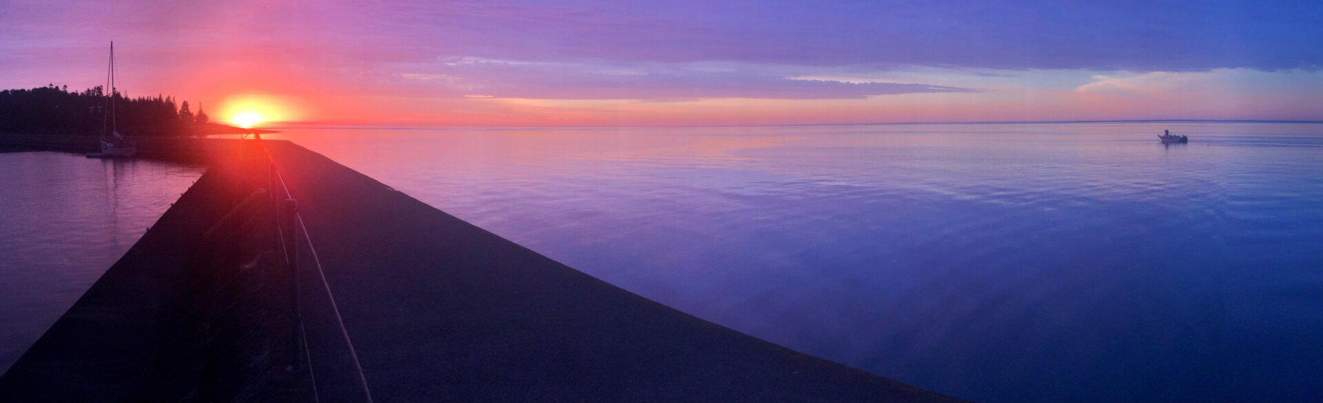 Two Harbors (N shore of Lake Superior) at dawn. A lone sailboat is moored next to the land-ward side of the breakwater. A lone fisherman is trolling out beyond the lake-ward side. The water is almost glass calm.