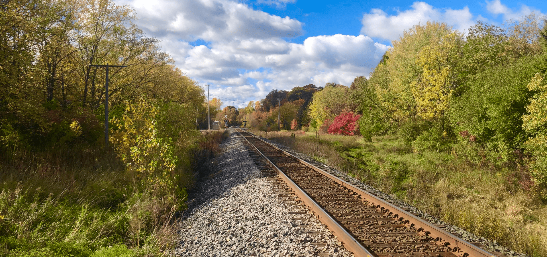 A sunny autumn Minnesota day, near noon, along a train track that extends from the bottom-right of the image up to about the middle, at which point it bends to the right in the distance. Deep blue sky 60% covered by puffy cotton-ball clouds. The leaves on either side of the tracks include deep and light greens, yellows, reds, & even a splotch or two of burgundy.