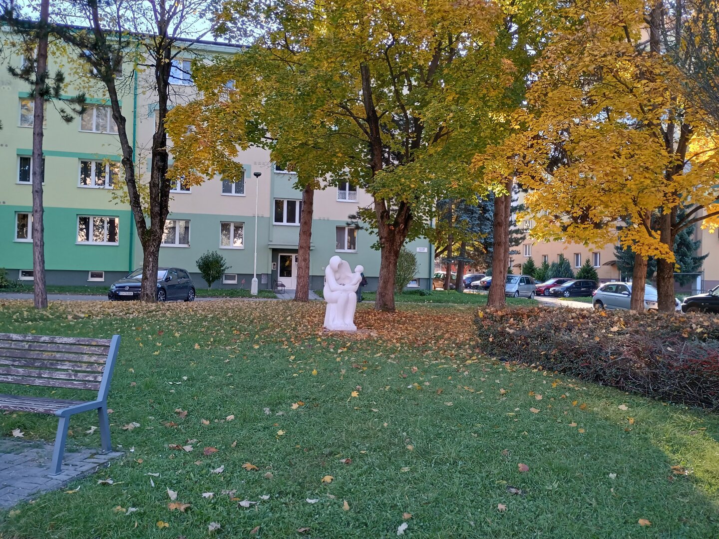 Socha v parku v pozadí sú stromy a dom napravo sú kríky naľavo je lavičk a v prostriedku socha matky s dieťaťom sediacim na jej nohách