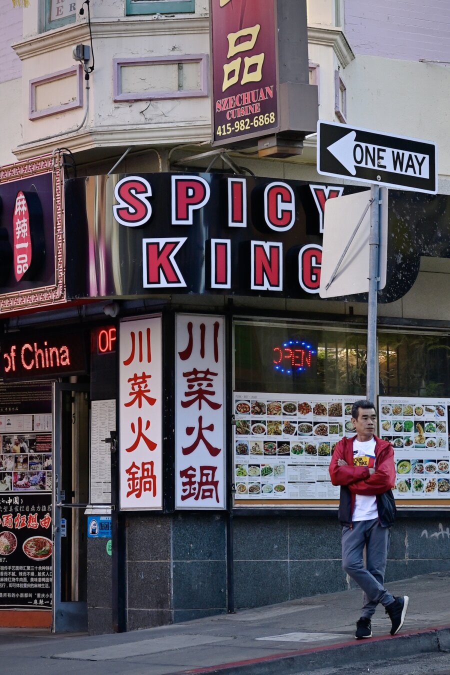 A man stands on the sidewalk, arms folded, right leg bent, right toes flexed. Red sleeves on his jacket match the red painted curb and the red signage of the hot pot restaurant behind him.