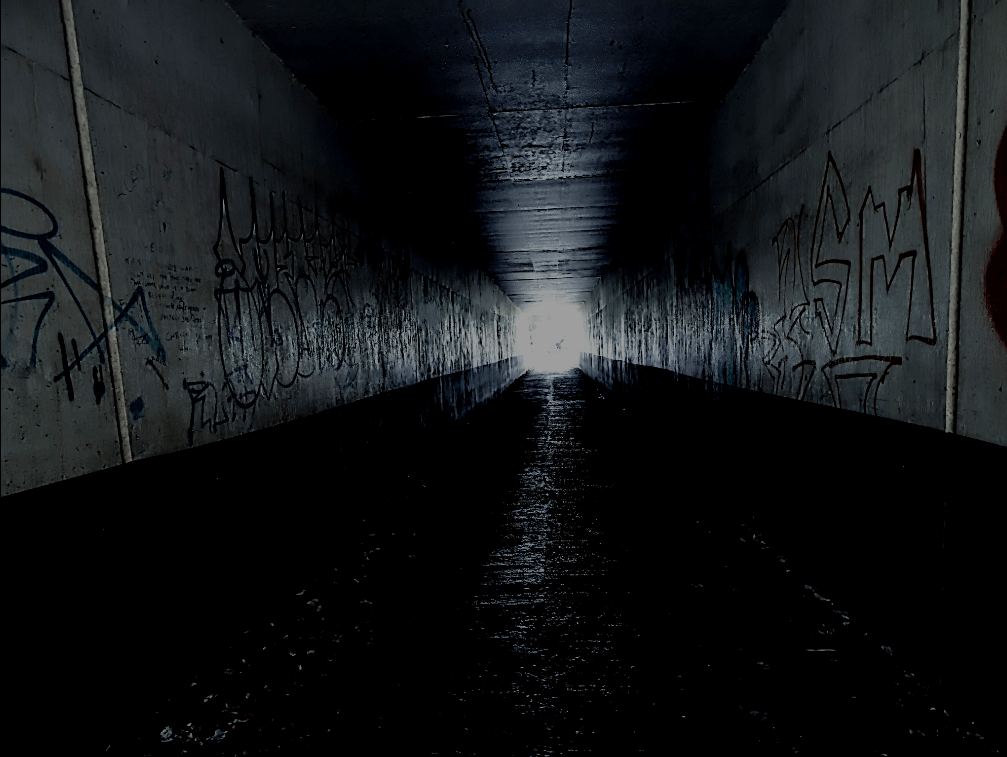 A pedestrian subway under a road . it's dark with a bright light at the end.