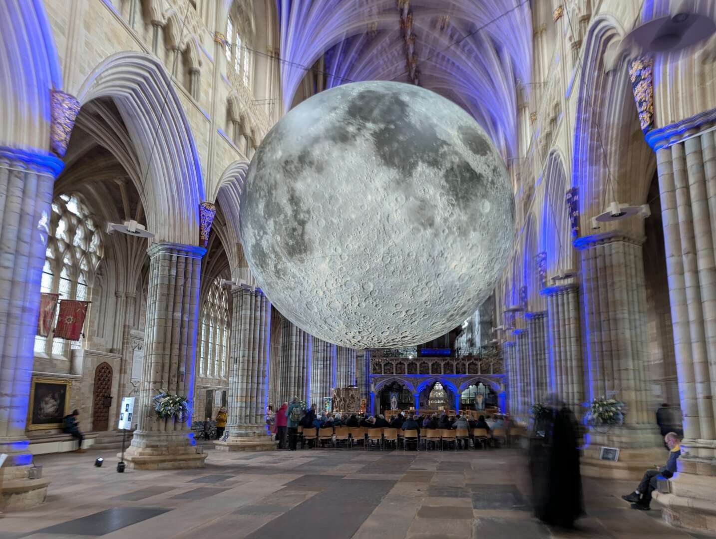 A sculpture of the moon hangs in the centre of the picture.  A ghostly figure of a priest is in the bottom right. The scene is set in a cathedral.