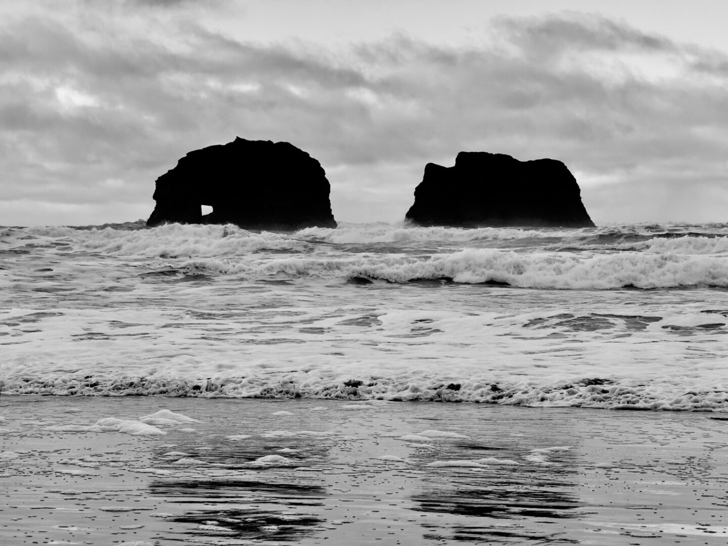 Two large rocks just off shore of a beach, the one on the left has an arch in it.