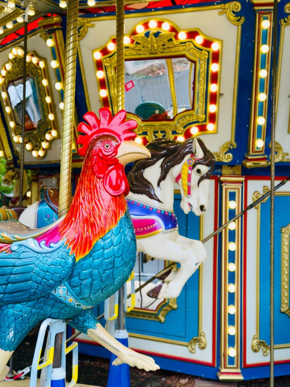 A colourful rooster (blue body, red head) merry go round figure.  There is a horse behind it and a mirror on the centre part of the merry-go-round.