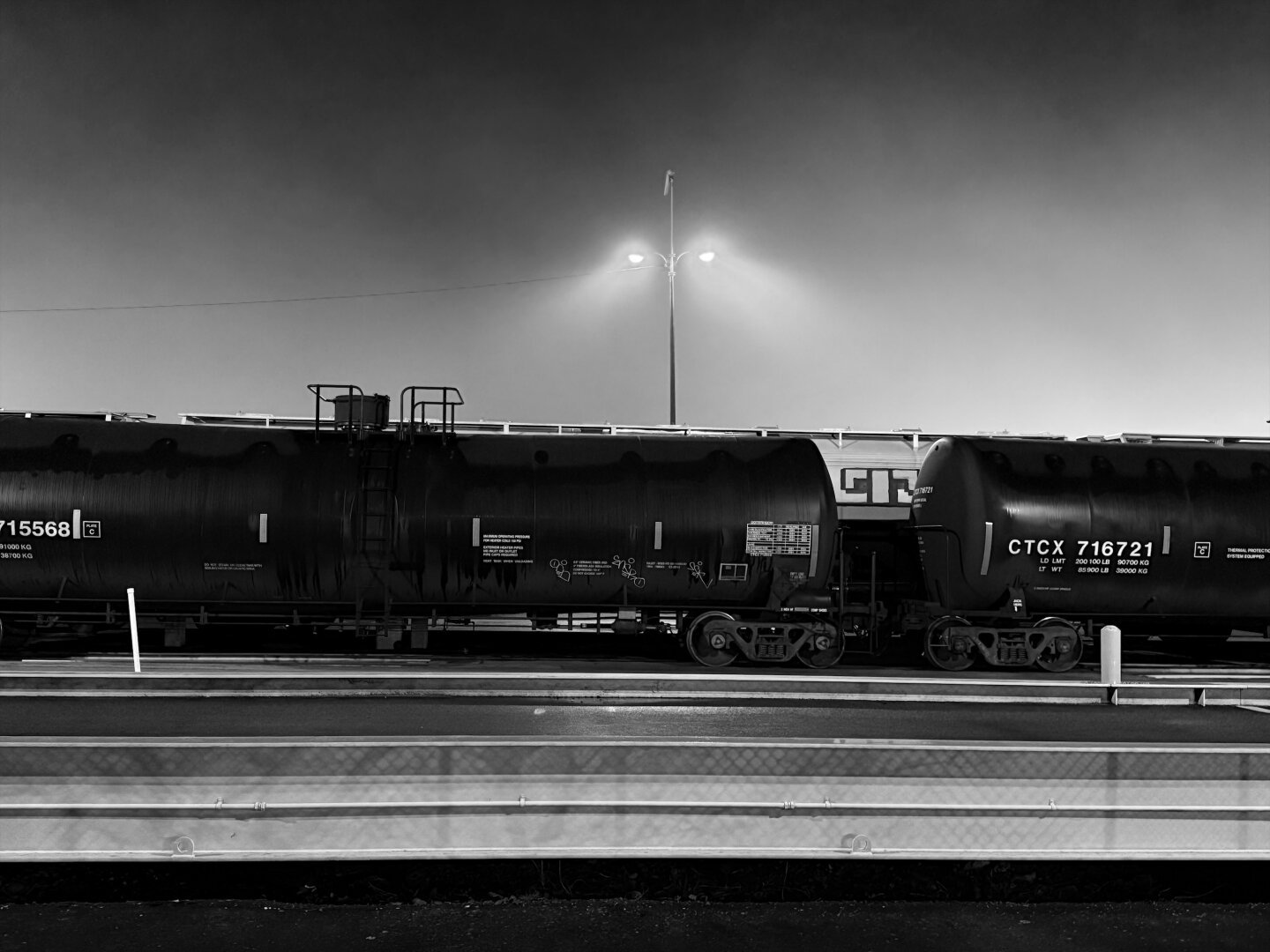 Black and white image of tanker cars in a rail yard.  There is a lamp post in the background with two lights, the light from the lamps is diffuse through some fog