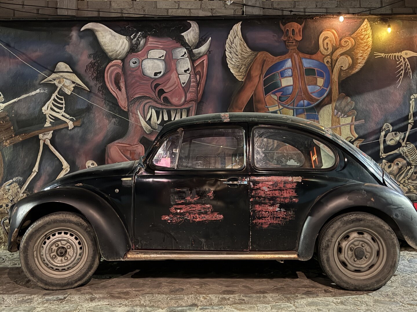 A black classic VW bug with some red marks on the side parked in front of a mural with pictures of devils.