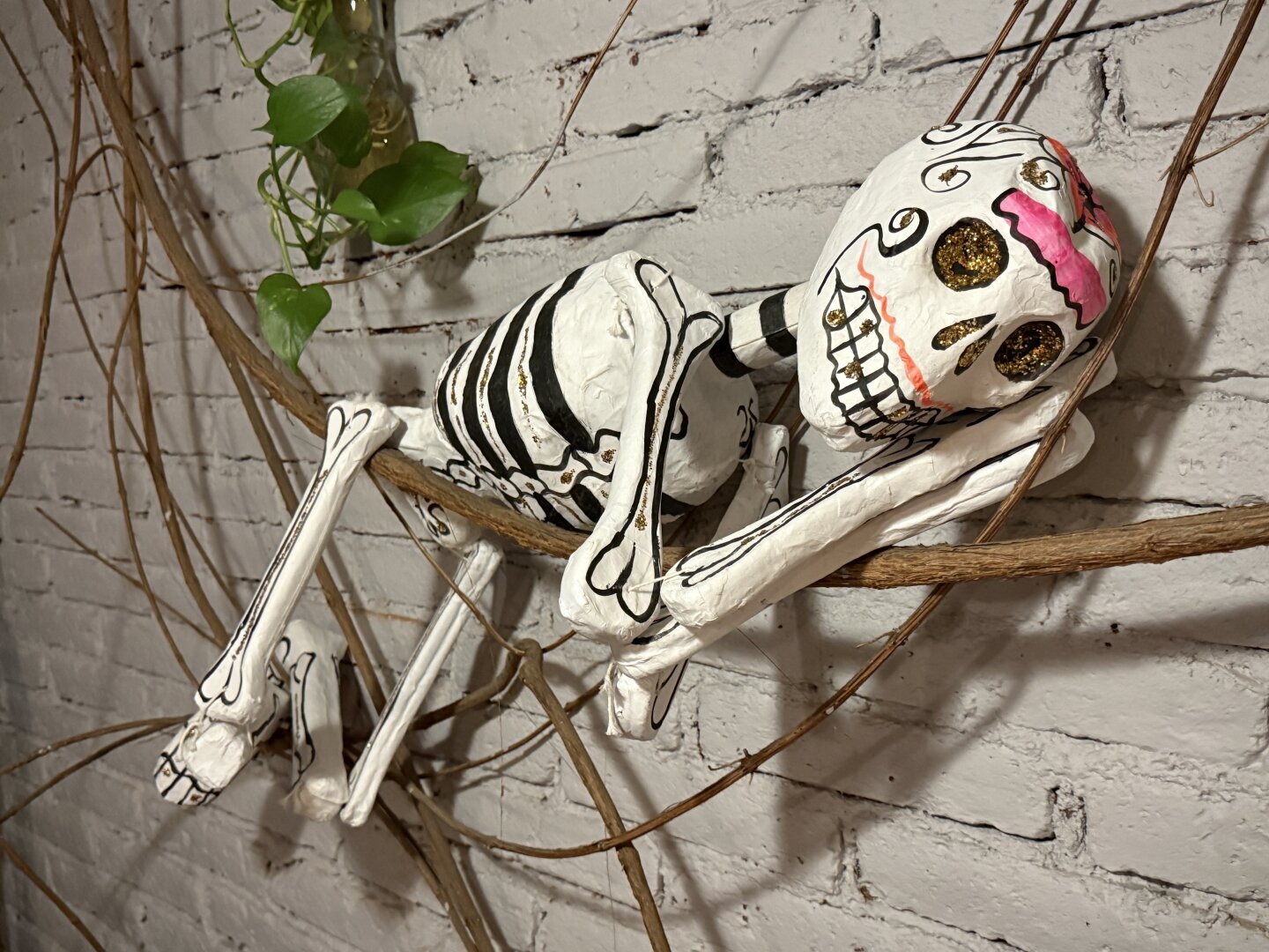 A small white skeleton painted with black stripes for ribs and mouth and decorated skull hanging on a vine in front of a brick wall. It has its hands folded under its head like it is sleeping.