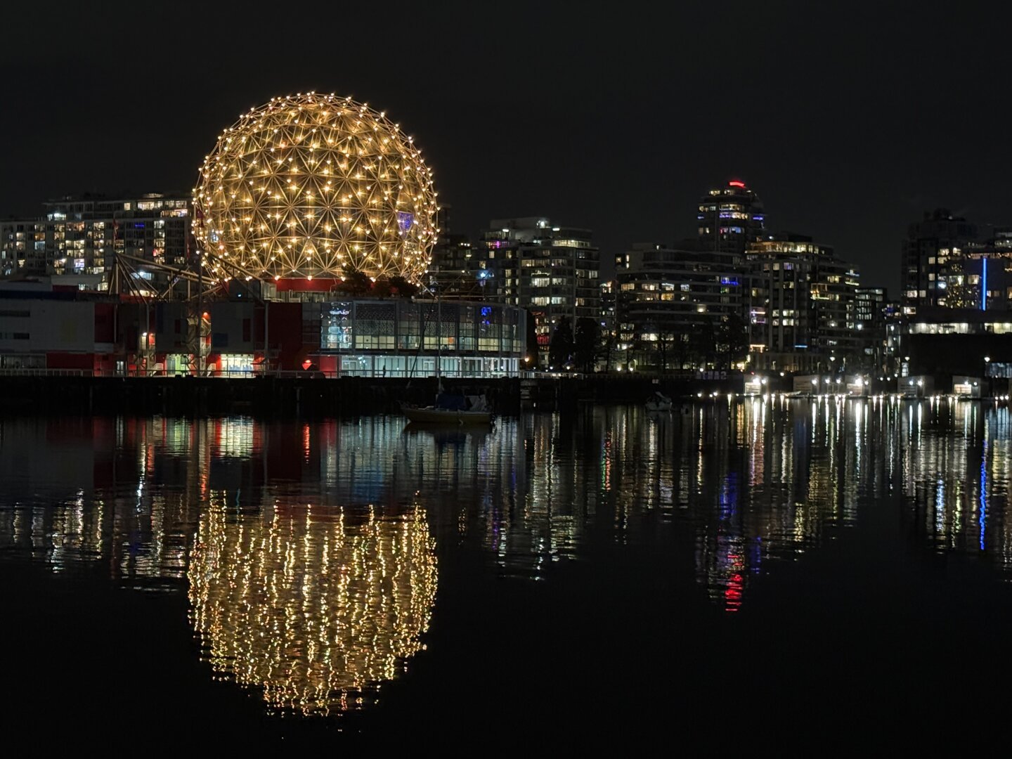 A geodesic dome at night with yellow lights. It is reflected in the water in the foreground. There are other buildings in the background and to the right.