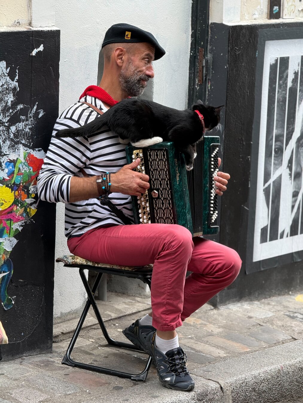 A man sitting on a folding stool wearing red pants, a black and white striped shirt with red collar or bandanna and a jaunty beret. He is playing the accordion and has a black cat with white feet and a red collar sitting on his accordion. What you can’t hear are the very stereotypically French songs he is playing.