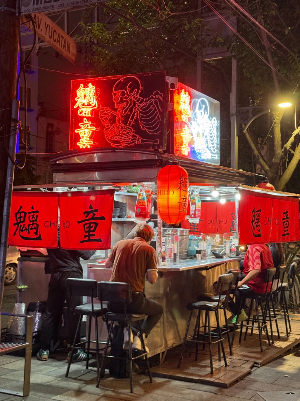 A street food stall serving ramen. There are red lanterns and flags with writing in Chinese and a bright neon sign on top showing a skeleton eating ramen out of bowl with chopsticks. There are a few customers seated around the food stall.