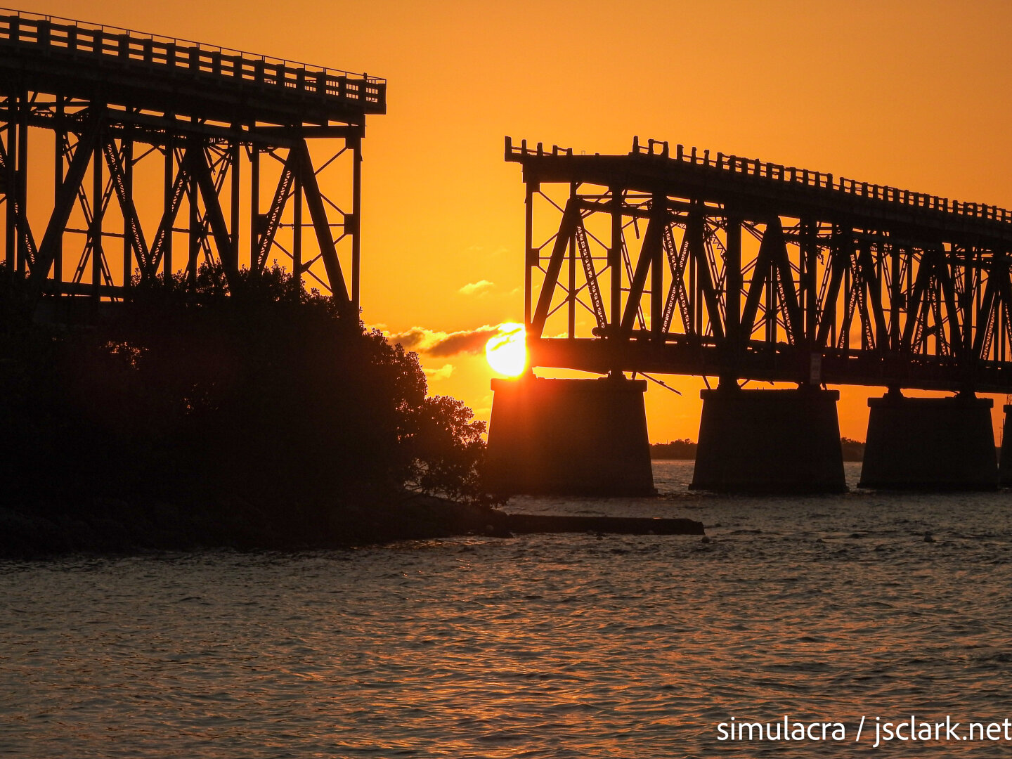 Orange sky and sunset behind an old truss railroad bridge.