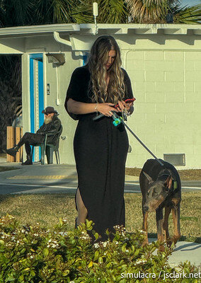 Candid shot: grizzled man in leather on a chair behind a nattily dressed woman staring at her phone while she walks her Doberman.