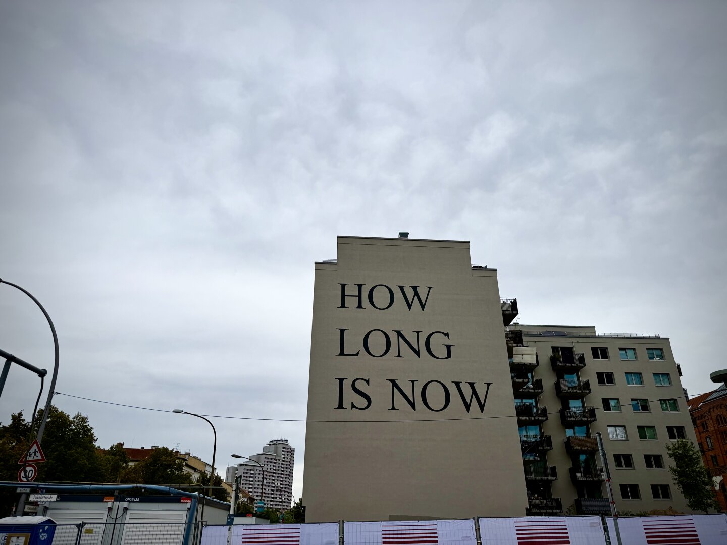A large windowless house features the text HOW LONG IS NOW in bold letters. The building is surrounded by urban elements, including street signs & construction barriers, under a cloudy sky