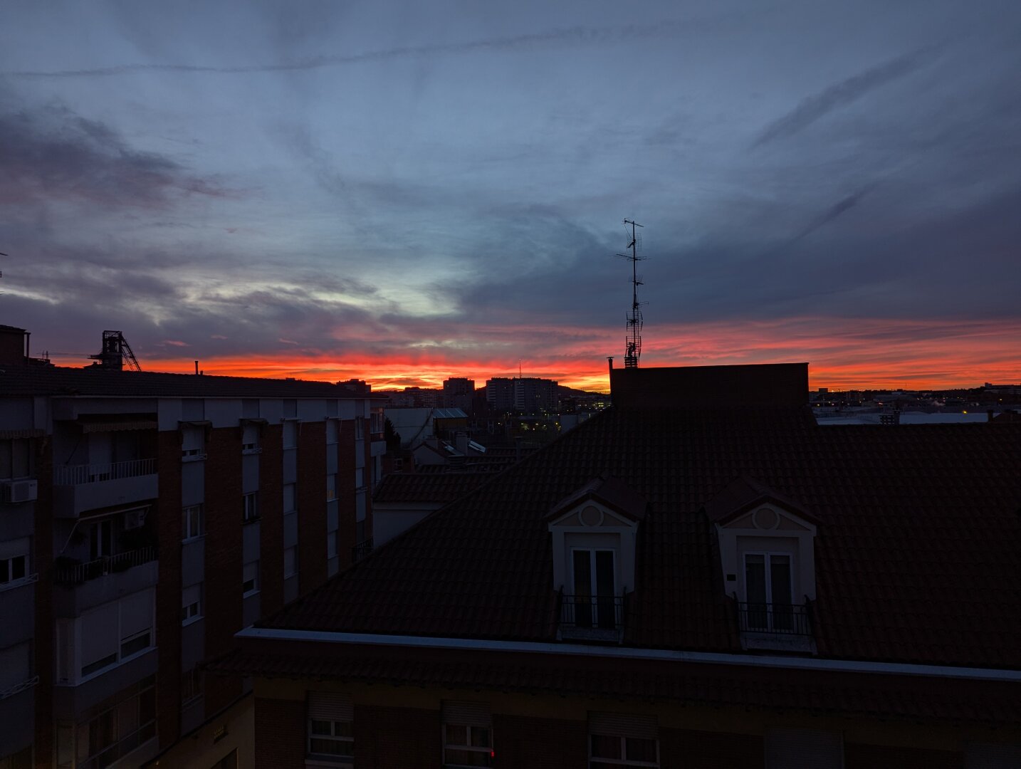 Siluetas de tejados de varias casas, casi indistinguibles en la primera luz del amanecer. Lo que más llama la atención es la línea roja en el horizonte, porque está saliendo el sol. El cielo tiene color plomizo en el resto de la vista.

También se recorta la silueta de una antena de televisión alta sobre el tejado de una casa.