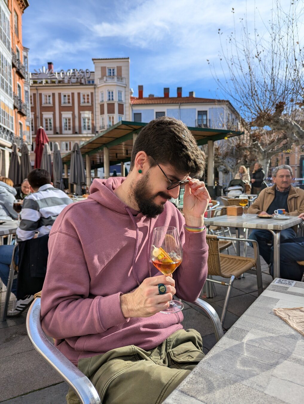 Andrés sentado en la mesa de una terraza. Viste una sudadera con capucha color rosa pastel y pantalones cargo verdes. Lleva un anillo y un pendiente verdes.

El día es soleado. Sujeta en su mano una copa de vermú. Está de perfil, mirando hacia un lado y sujetándose sus gafas de sol como si fuese a quitárselas en el siguiente instante.