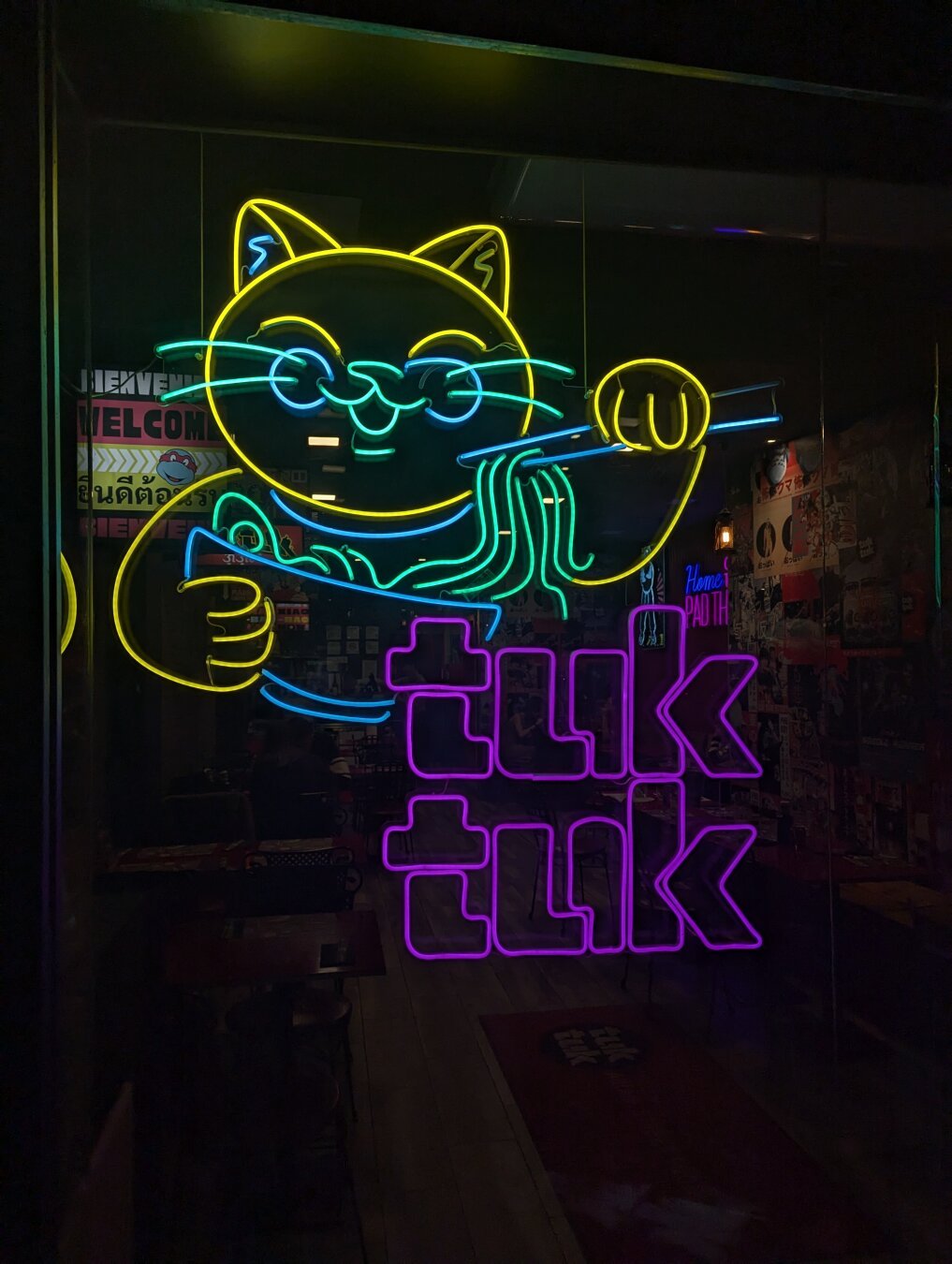 Un letrero luminoso en un cristal. Representa a un gato comiendo fideos de un plato hondo, con palillos. Junto a él pone "Tuk tuk". El gato es de color amarillo y verde neón, y las letras rosas. Es un cartel muy llamativo porque además la foto se tomó de noche.