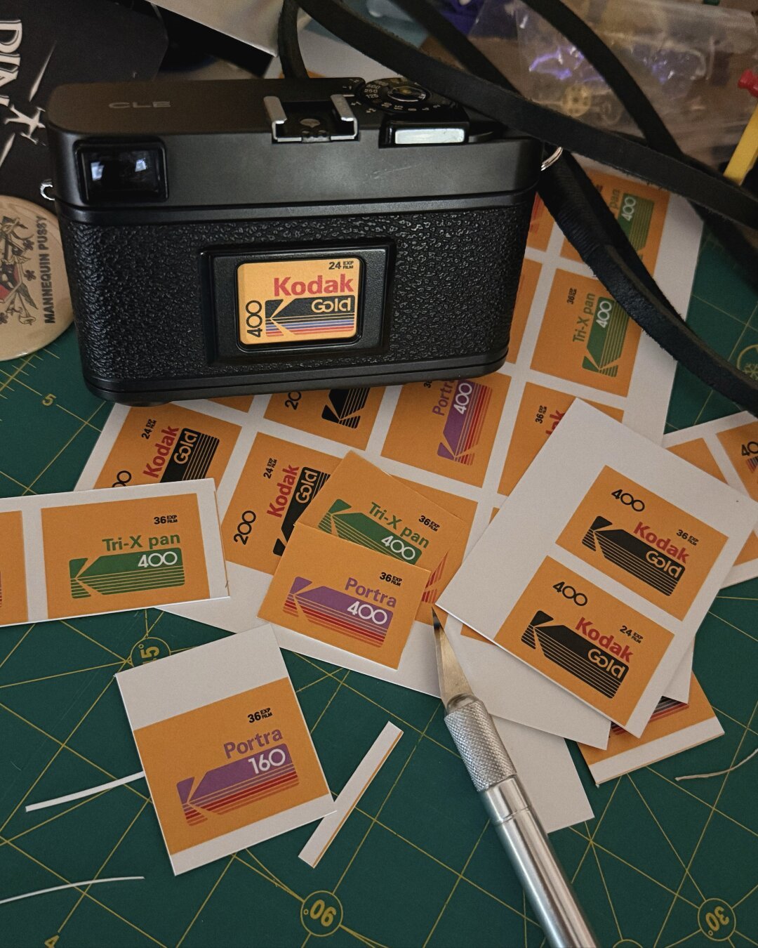 A Minolta CLE camera with a custom film box tab in the memo slot on the back on a pile of various film box tabs for Kodak Portra, Kodak Gold, Kodak Tri-X, etc.