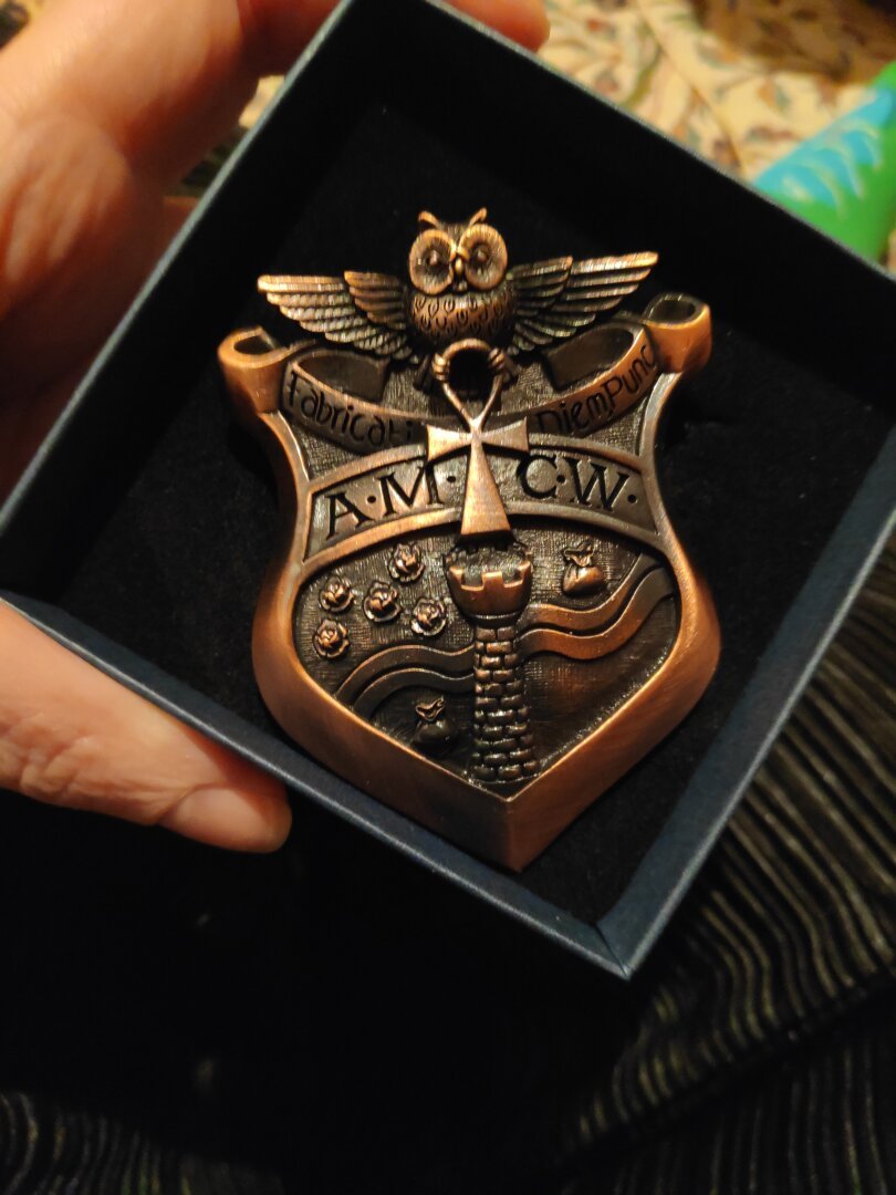 Authentique badge d'agent du Guet de Ankh-Morpokh dans son écrin ! 
Il a la forme du blason de la ville, traversé par un bandeau avec les initiales AMCW (guet de la ville de Ankh Morpokh)
Le blason : une tour au centre, crénelée a son sommet. Derrière, en biais, une rivière un tracé onduleux. La tour et la rivière séparent le fond en 4 quartiers. Quartier en haut à gauche : 5 choux. Quartier en haut a droite : une bourse. Quartier en bas a droite : une bourse. Quartier en bas a gauche : vide. La chouette a l'Ankh domine le blason.