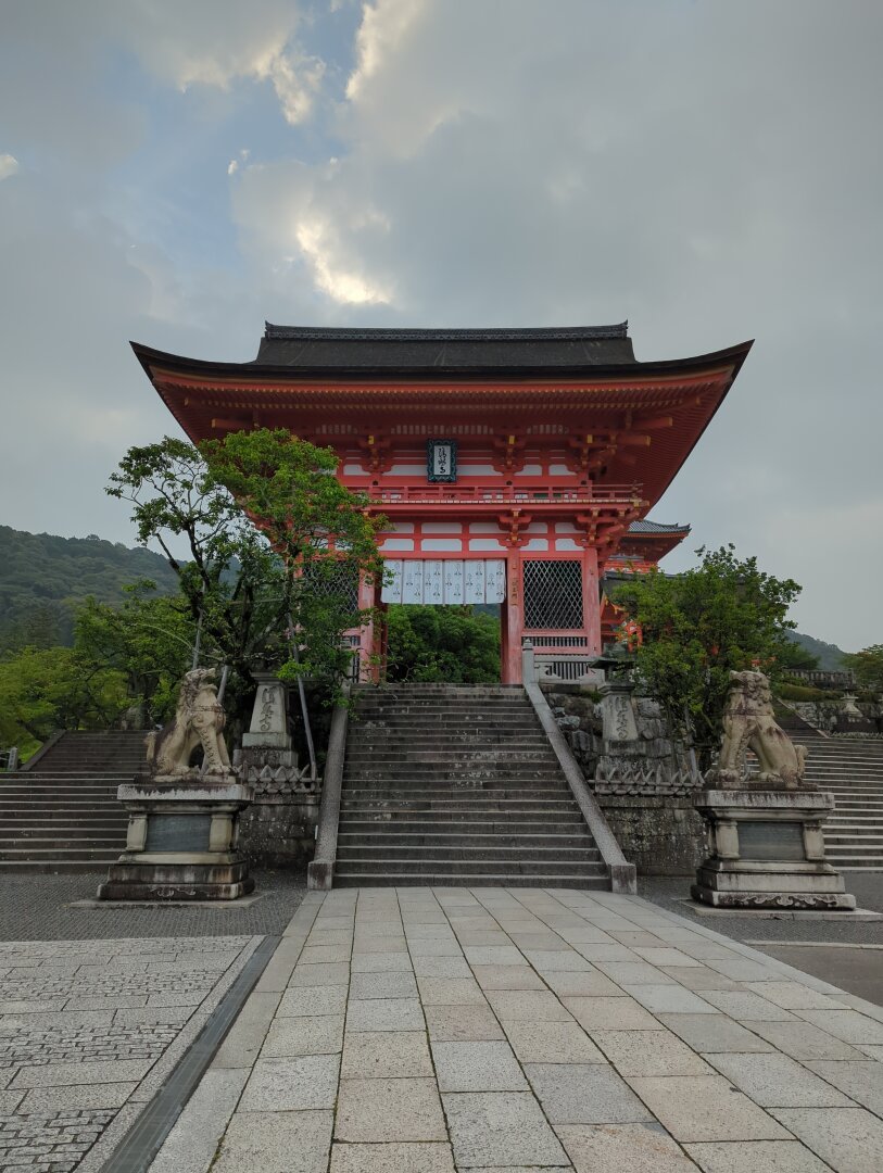 Front stairs and entrace gate of Kyomizudera in Kyoto. Flanked on either side by komainu (lion-dog) statue guardians. Background of rolling green Kyoto mountains.