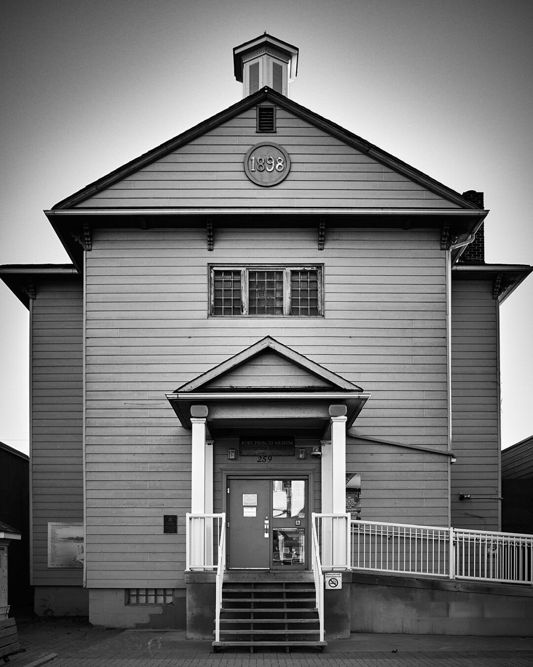 The image shows a building with a peaked roof, featuring a sign that reads '1898' at the top. The front entrance has a set of stairs leading up to a door labeled 'FORT FRANCIS MUSEUM' with a number '259' above it. The building is painted in a light color and has multiple windows, some of which are partially obscured.