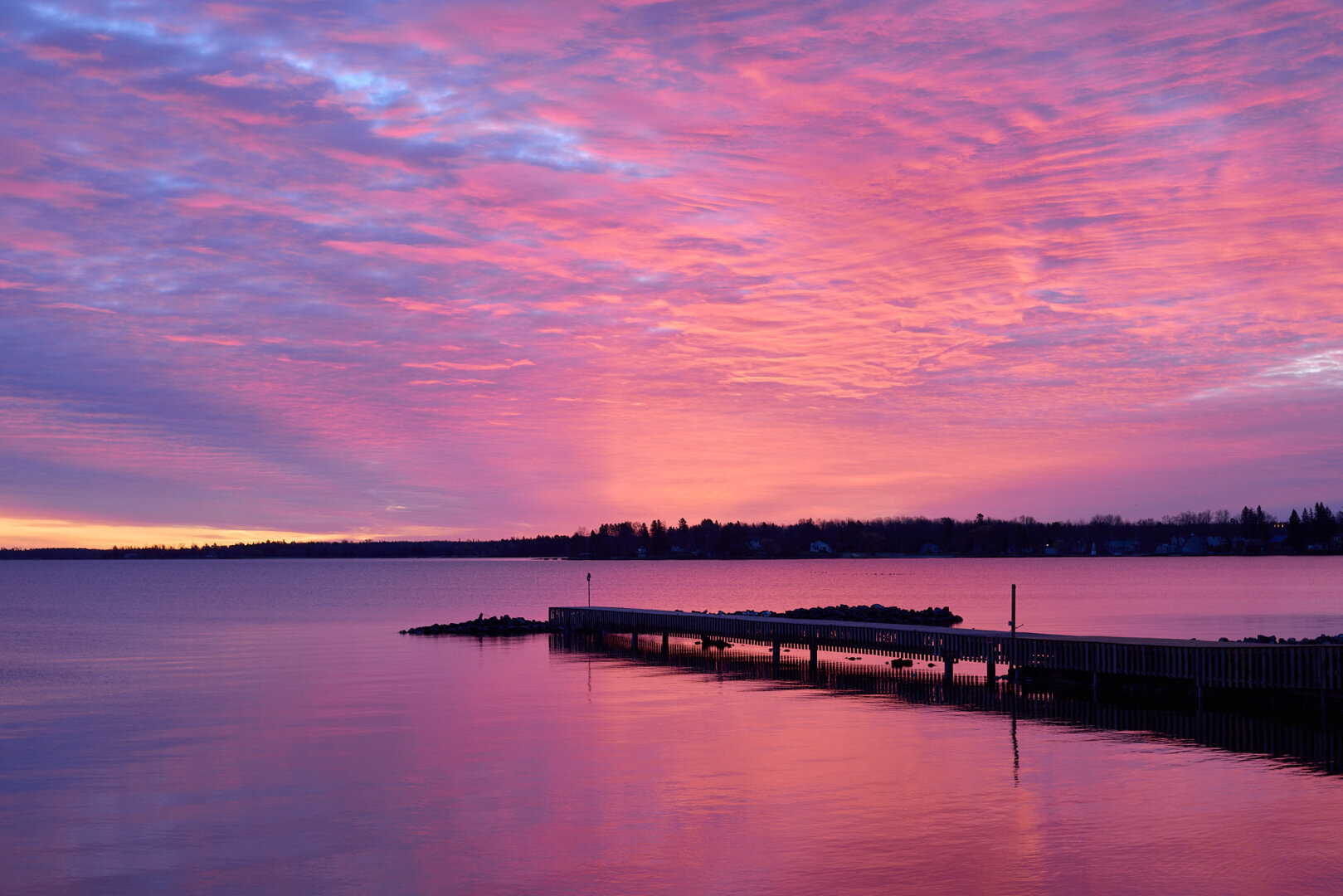 A tranquil lake scene at sunrise, featuring a wooden dock extending into the water. The sky is filled with vibrant pink and purple clouds reflecting on the calm surface of the lake. In the background, silhouettes of trees and buildings are visible along the shoreline.