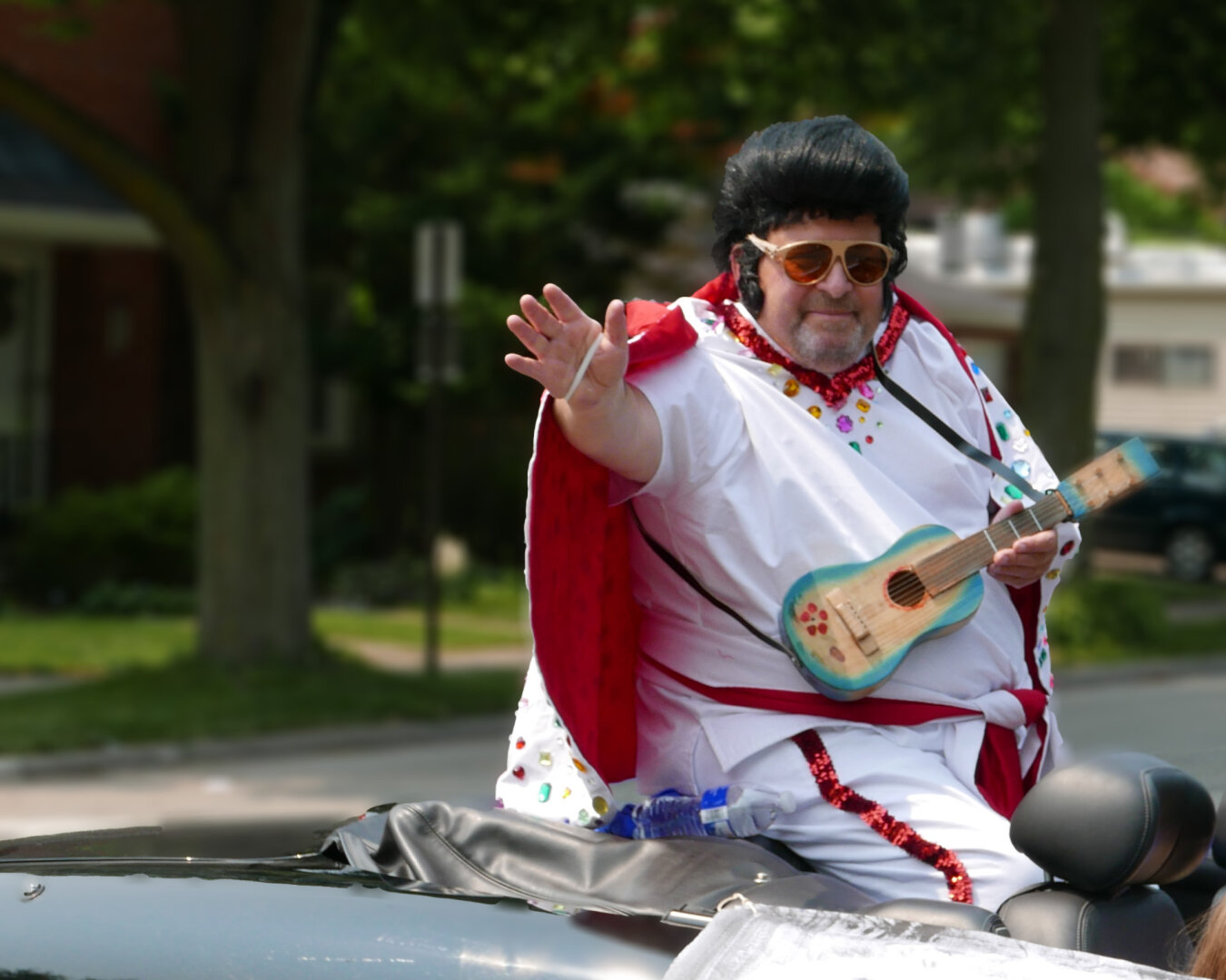 A person wearing a white outfit with colorful embellishments and a red cape is seated on a car. They have dark hair styled in a pompadour and are holding a small blue ukulele. The background features trees and a residential area, with a street sign partially visible.