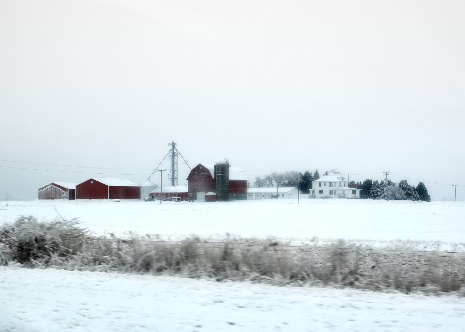 The image shows a snow-covered landscape with a red barn and silos on a farm. In the distance, there is a white house surrounded by frost-tipped trees. The sky is overcast, casting a soft, muted light over the scene, emphasizing the cold, tranquil atmosphere.