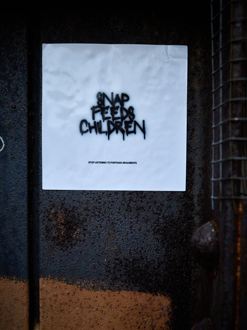 A white poster with bold black lettering stating “SNAP FEEDS CHILDREN” is attached to a rusted metal surface beneath a bridge. The metal around it is dark, rough, and weathered, emphasizing the harsh setting.