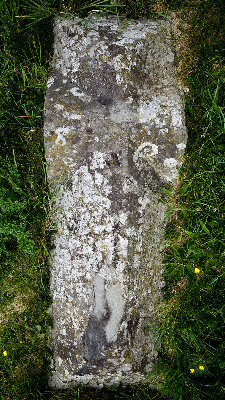 A weathered grave stone lies flat on the ground, partially covered by grass. The stone has a rough texture with patches of lichen and moss. It is surrounded by green grass and a few small yellow flowers.