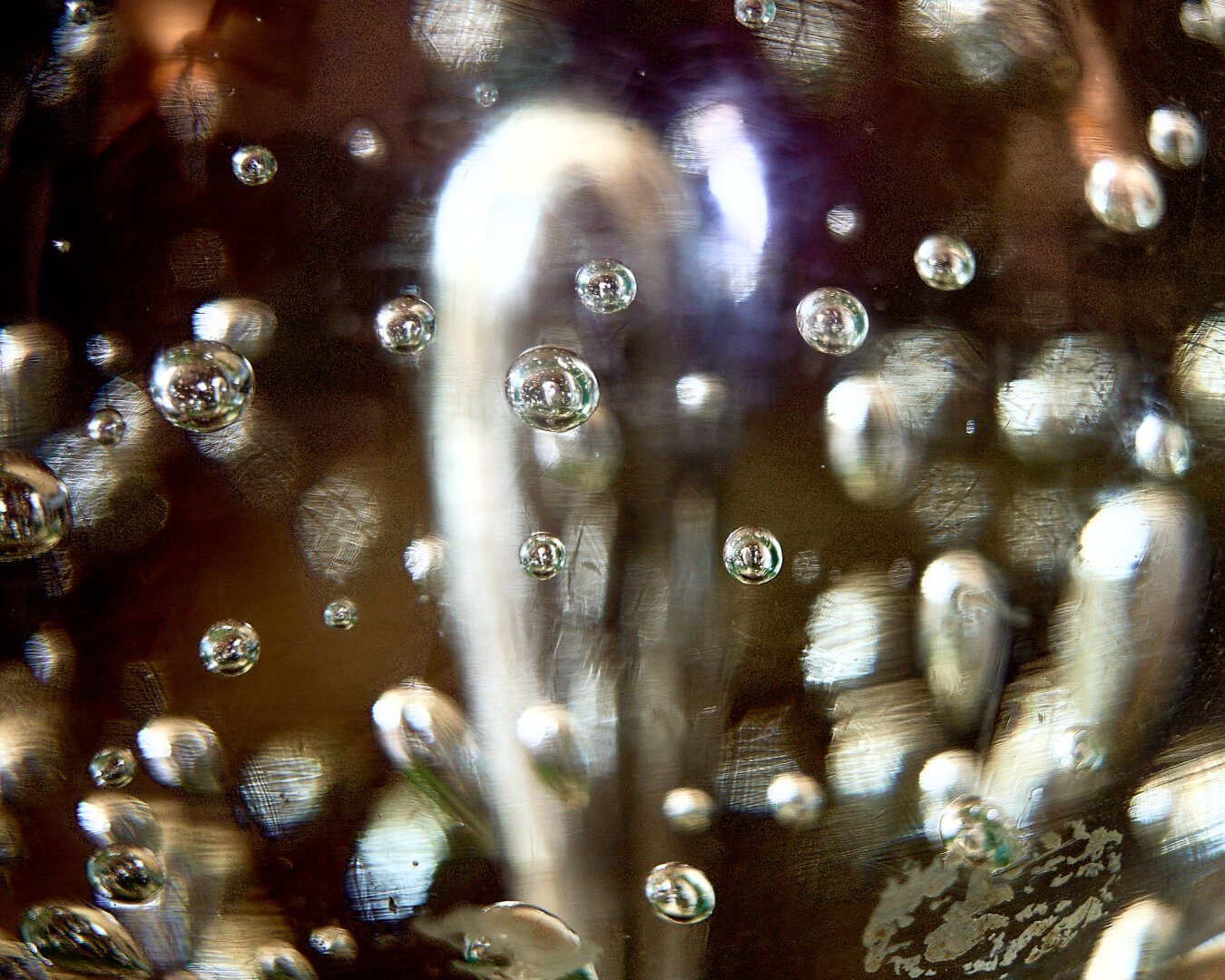 A close-up view of a glass object filled with bubbles. The glass surface is reflective, showcasing multiple spherical bubbles of varying sizes. The background appears blurred with light reflections, creating a shimmering effect.
