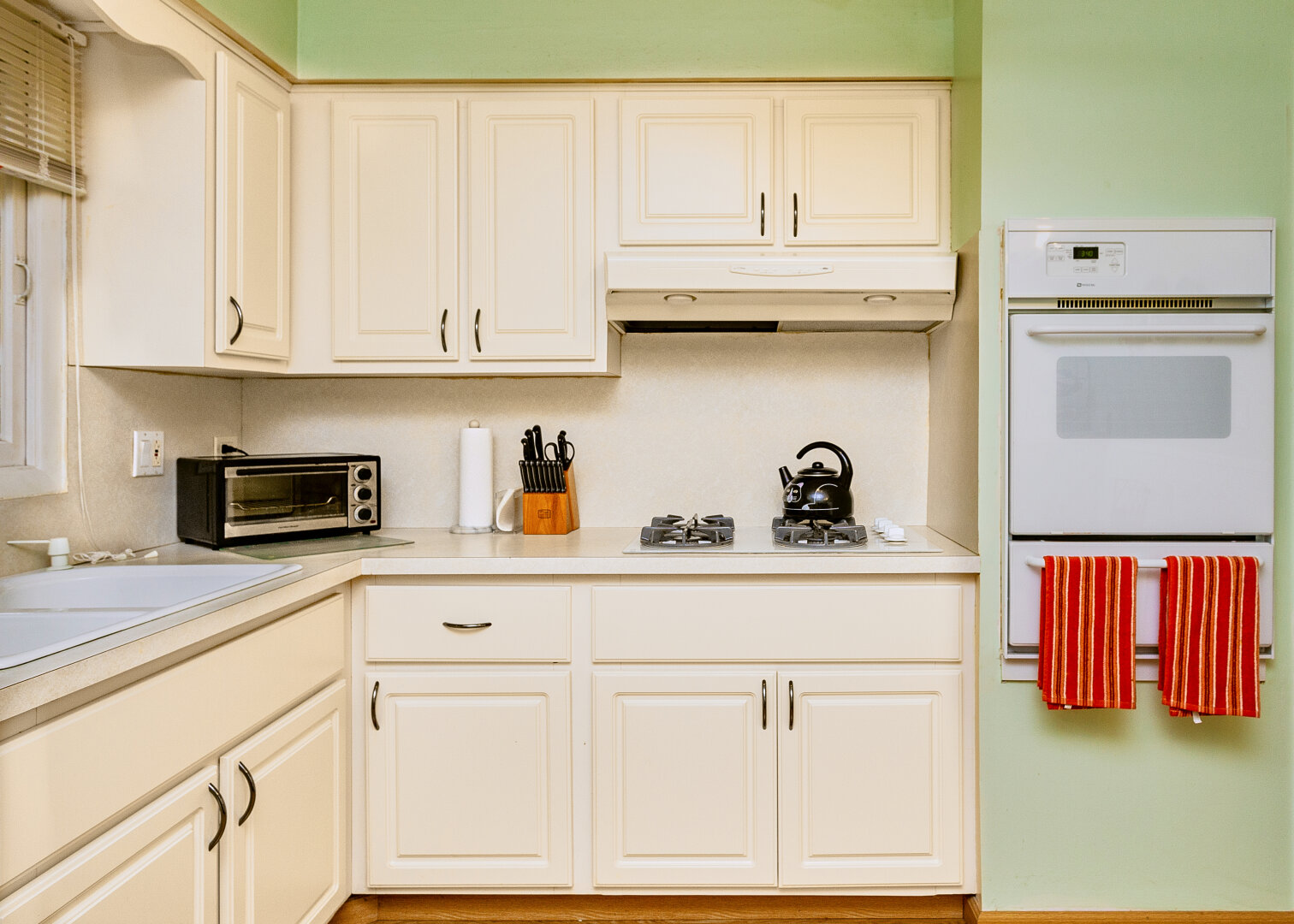 A kitchen with light-colored cabinets and a green wall. On the countertop, there is a black microwave, a white paper towel holder, and a wooden block with knives. A black kettle sits on a gas stove with two burners. To the right, there is a white oven with two red-striped towels hanging from it.